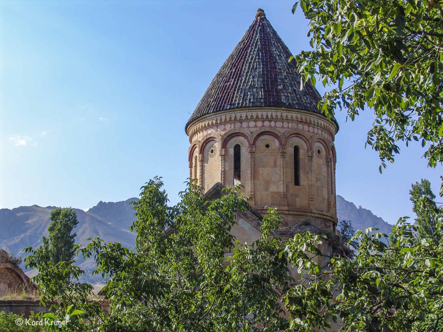 Ein reich verzierter steinerner Turm mit kegelförmigem Dach erhebt sich zwischen grünen Bäumen vor einer kargen Bergkette.