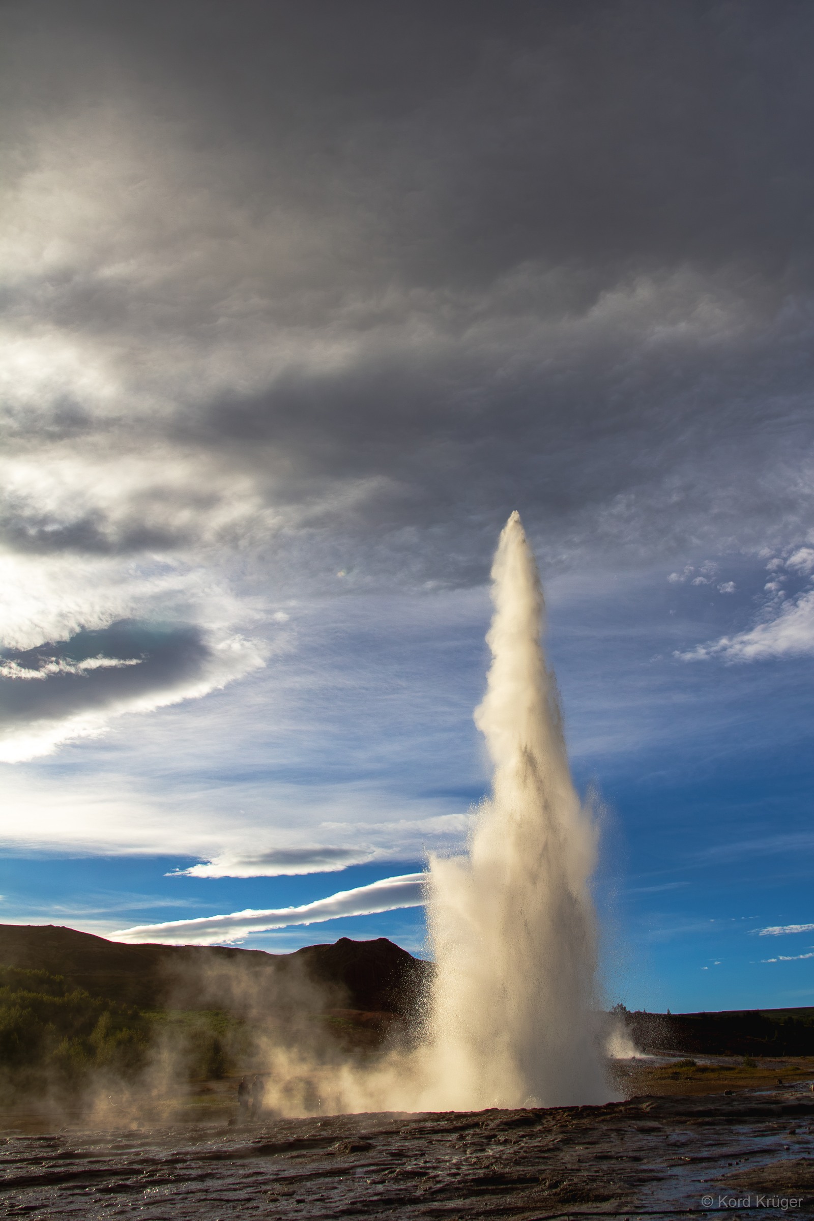 Eine hohe helle Wasserfontäne steigt aus einer dunklen Geothermalfläche vor einem wolkigen Himmel auf.