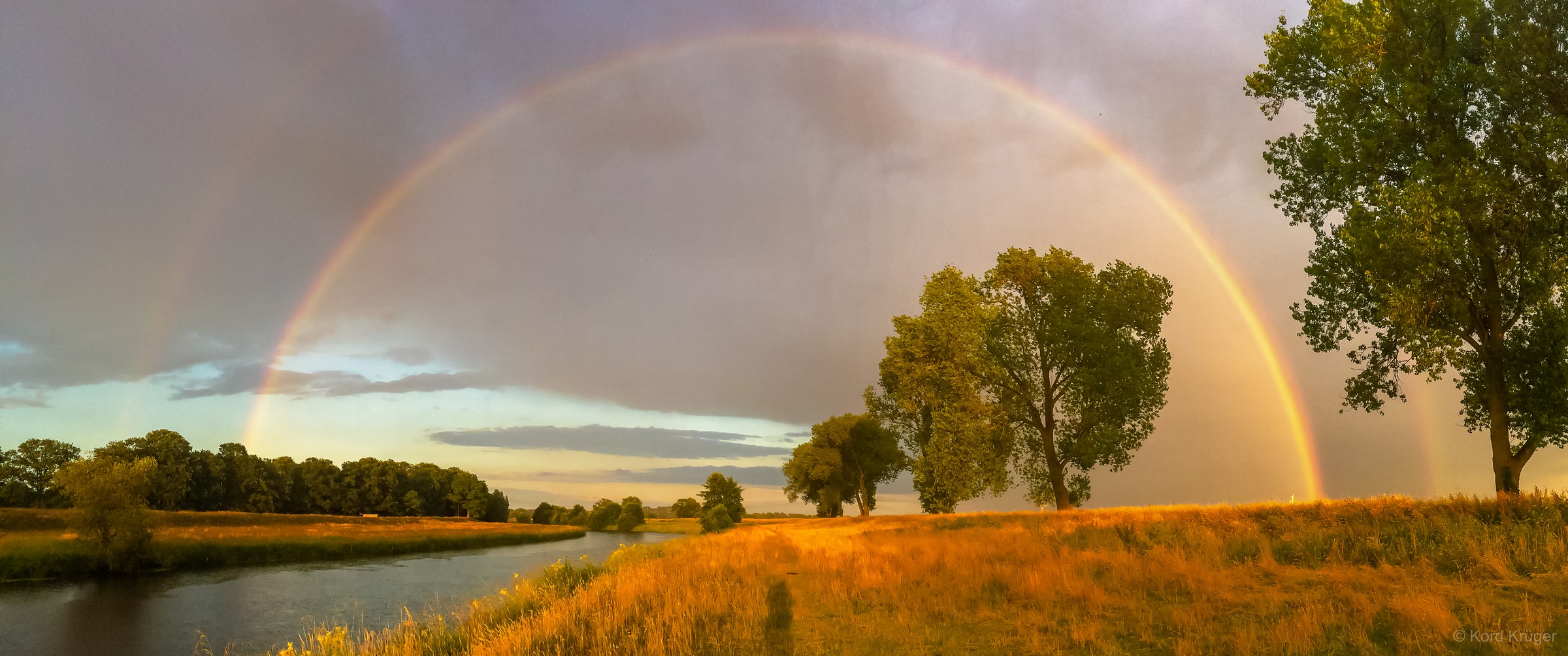 Ein Regenbogen spannt sich über einen Fluss und eine von Bäumen gesäumte Uferlandschaft im warmen Licht.