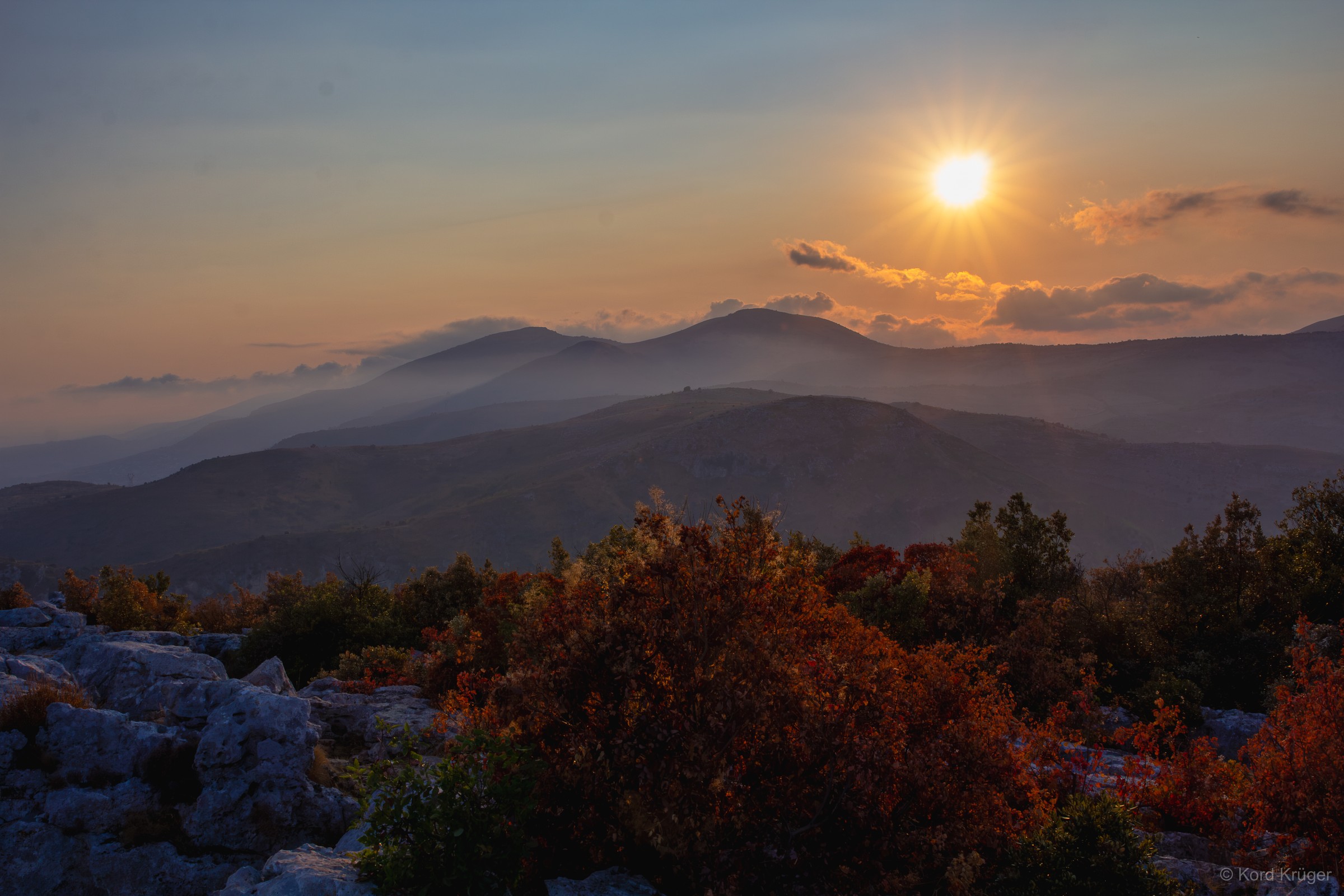Die tief stehende Sonne leuchtet über mehreren blaugrauen Bergketten; Sträucher rahmen den Vordergrund.