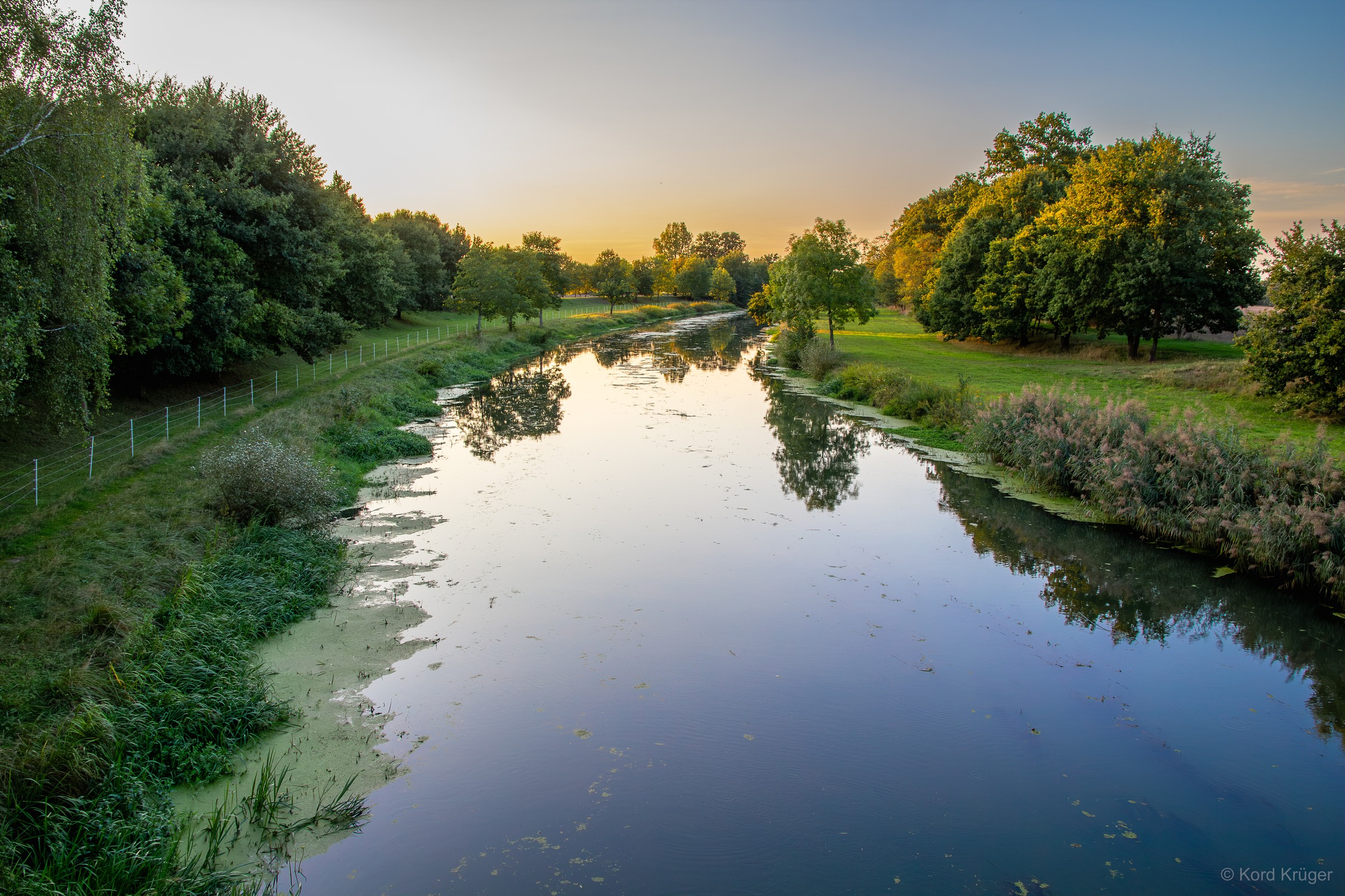 Ein ruhiger Fluss führt zwischen grünen Ufern und Bäumen in die Ferne und spiegelt den hellen Himmel.