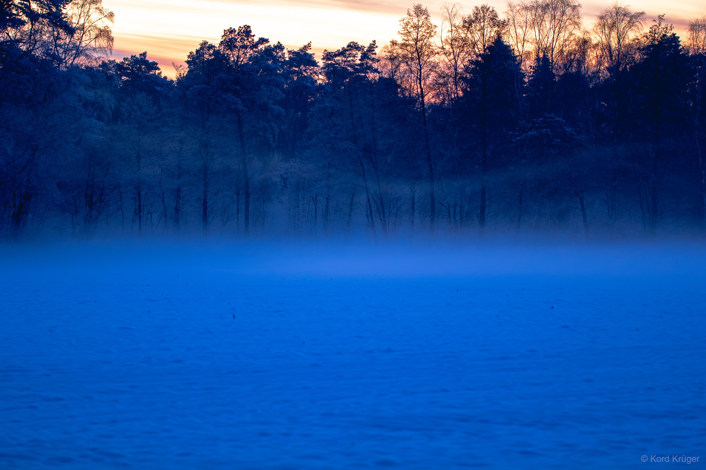 Blauer Dunst liegt über einer winterlichen Wasserfläche vor dunklen Bäumen und einem schmalen warmen Himmelsstreifen.