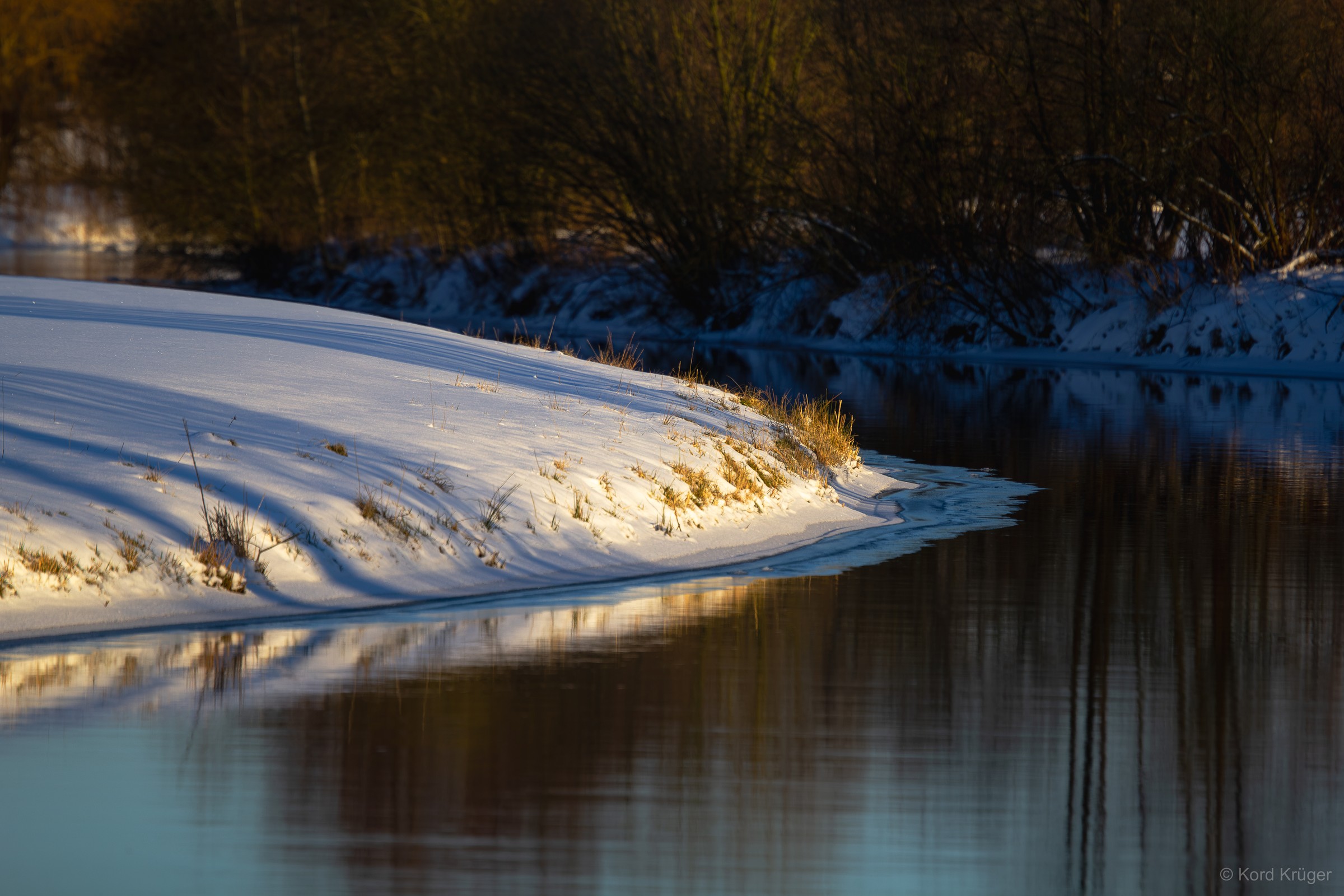 Ein verschneites Ufer mit Bäumen und trockenem Gras spiegelt sich in dunklem, ruhigem Wasser.