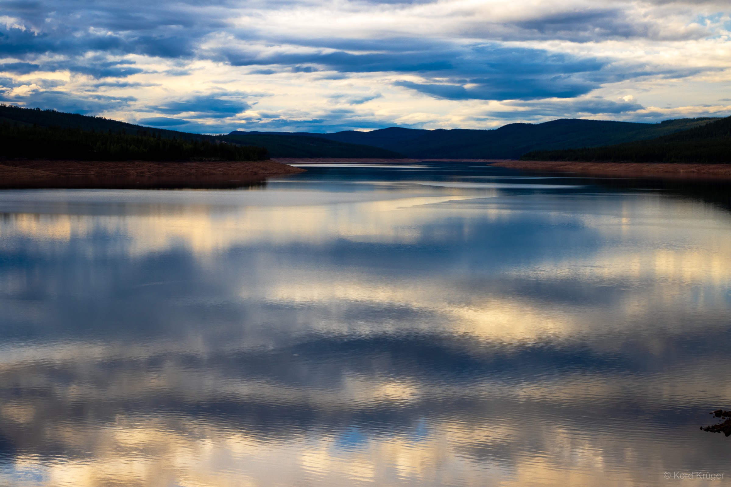 Ein ruhiger See spiegelt graublaue Wolken vor einer dunklen Bergsilhouette und einem flachen Ufer.
