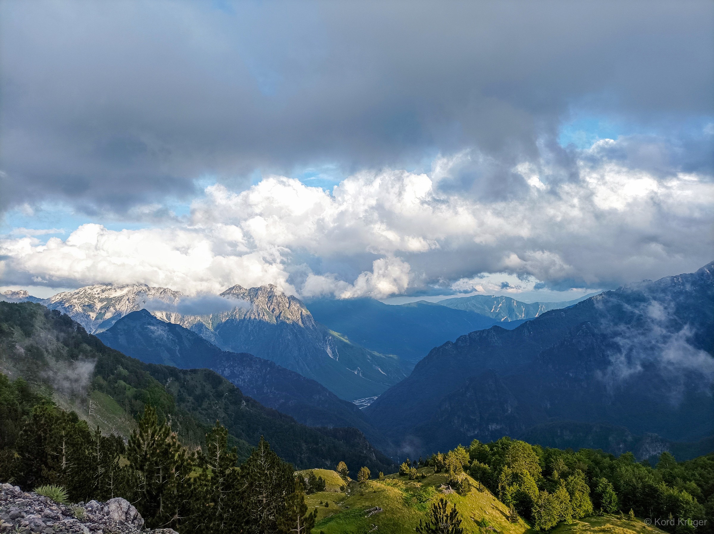 Mehrere grüne und blaue Bergzüge staffeln sich unter schweren grauweißen Wolken um ein tiefes Tal.
